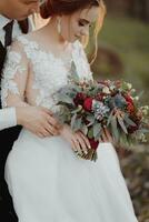 Photo of a wedding bouquet of white different and greenery in the hands of the bride and groom. The groom holds the bride's hand. Beautiful hands. French manicure. Wedding rings