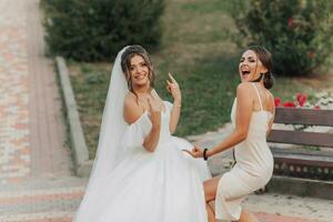 Wedding photo in nature. A brunette bride in a white long dress and her friend in a nude dress are fooling around together and smiling sincerely. Young women. emotions