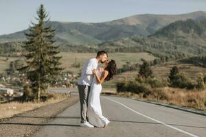 A stylish groom in a white shirt and a cute brunette bride in a white dress are hugging and kissing on an asphalt road against the background of a forest and mountains. Wedding portrait of newlyweds. photo