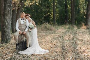 Wedding photo in nature. The groom is sitting on a wooden stand, the bride is holding a bouquet, standing next to him, leaning on his shoulder. Portrait of the bride and groom