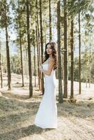 A girl with long black curly hair blowing in the wind is standing in a white silk dress. Standing in the spring forest against the background of large trees photo