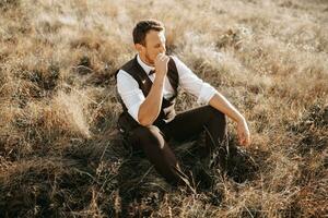 portrait of a stylish groom on a background of dry autumn grass. the concept of a rural wedding in the mountains, happy bohemian newlyweds. man relaxing sitting on the grass photo