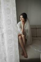 Portrait of the bride. A brunette bride is sitting on a nude sofa in a robe and a long lace veil, posing. Gorgeous make-up and hair. Wedding photo. Beautiful bride photo