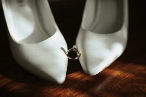 Photo of details at the wedding. The wedding ring of the bride stands on white classic shoes, which are located on a wooden background. Contour line. Shadows and light.