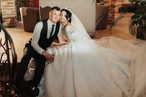 Wedding portrait of the bride and groom. Indoor photography with beautiful light. Stylish Groom in a black vest kisses a brunette bride on the cheek, sitting hugging indoors. Couple in love photo