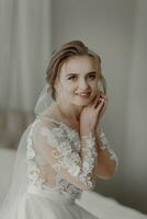 Preparation for the wedding. Beautiful young bride in white wedding dress indoors. Luxury model looking out window at home in studio room with big window. The girl shows photo
