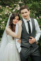 Happy young couple standing near beautiful flowers. The bride gently hugged her groom behind the shoulders. Young people are gently hugging, looking at the camera. Wedding portrait. Spring wedding photo