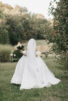 Wedding photo. The bride in a voluminous white dress and a long veil walks in the garden along a stone path, holding a bouquet of white roses, her shoulders are turned to the camera. photo