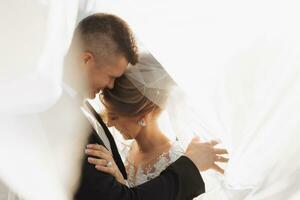 Wedding portrait. The groom in a black suit and the blonde bride are hugging, wrapped in the bride's veil. Long dress in the air. Photo session in nature. Beautiful hair and makeup