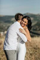 A stylish model couple in the mountains in the summer. A young boy and a girl in a white silk dress are walking on the slope against the background of the forest and mountain peaks. boy kisses a girl photo