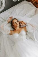 A beautiful curly brown haired bride in a white dress poses for a photographer while lying on a bed in a beautiful dress with sleeves. Wedding photography, close-up portrait, chic hairstyle. photo