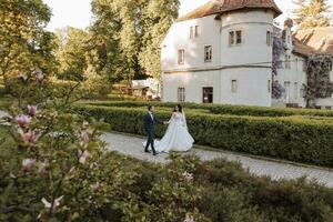 contento joven pareja, novia con largo Rizado pelo en blanco vestir con largo tren cerca castillo en hermosa flores hermosa niña en el parque. hermosa luz de sol. Boda foto disparar