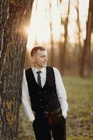 Portrait of the groom. The groom in a black vest is standing in the forest, leaning on a tree and touching his chin, looking away. Wedding in nature photo