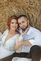 Wedding portrait. The bride and groom are sitting in an embrace on the ground, near a bale of hay and looking into the lens. Red-haired bride in a long dress. Stylish groom. Summer. photo