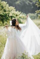 A beautiful bride with long curly hair holds her delicate dress and veil and admires it in the garden. Morning of the bride. Natural makeup. Good daylight. photo