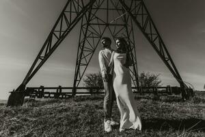 Stylish model couple in the mountains in summer. A young boy and girl in a white silk dress are standing near large structures of power lines. Black and white contrast photo