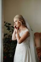 hermosa joven novia vistiendo pendientes antes de Boda ceremonia a hogar. un rubia novia en un Boda vestir usa pendientes foto