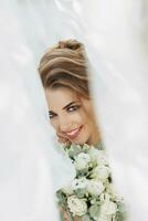 Curly blonde bride in a white dress, covered with a veil, poses for the camera with a bouquet of roses. Portrait of the bride. Beautiful makeup and hair. Wedding in nature photo