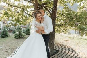 Wedding portrait. A groom in a black suit and a blonde bride are standing, hugging and kissing under a tree. Photo session in nature. A beautiful ray of the sun in the photo. Beautiful hair and makeup