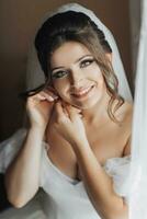 A brunette bride in a white dress puts on her earrings and smiles sincerely at the camera. Open shoulders. Beautiful hands. Long veil. Morning of the bride. photo