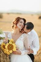 Wedding portrait of the bride and groom. The groom, tearing his shirt, stands behind the bride, near a bale of hay. Red-haired bride in a long dress with a bouquet of sunflowers. Stylish groom. Summer photo