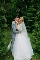 Portrait of the bride and groom standing on the background of green trees, embracing. The bride smiles sincerely. Stylish groom. Fashion and style. Beautiful bride photo
