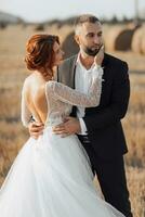 Boda retrato de novias y novios en pie abrazando el Mira de el novia y un amable toque a su rostro. Pelirrojo novia con abierto espalda. elegante novio. verano foto