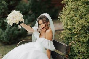 Portrait of the bride in nature. A brunette bride in a white long dress, holding a bouquet of white roses, poses, looking into the lens, smiling sincerely. on the background of the tree. Curly hair. photo