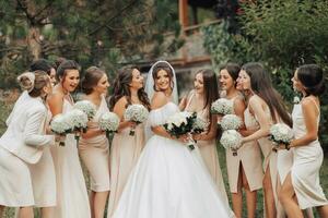 Wedding photo in nature. A brunette bride in a long white dress and her friends in nude dresses stand against a background of trees, smiling and holding their gypsophila bouquets. Young women.