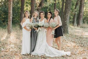 Wedding photo in nature. The bride and her bridesmaids are standing in the forest smiling, holding theirs bouquets and looking into the camera lens. Happy wedding concept. Emotions. girls