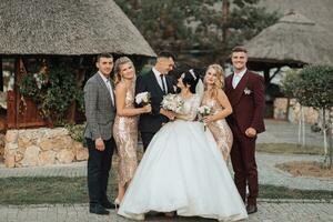 Front view of newlyweds standing and hugging, between friends. Girls in golden dresses hold bouquets, the bride in a voluminous dress, the groom and his friends in suits. Wedding in nature photo