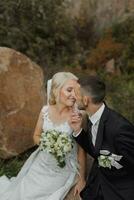 happy beautiful bride and elegant stylish groom sitting on a rock and kissing photo