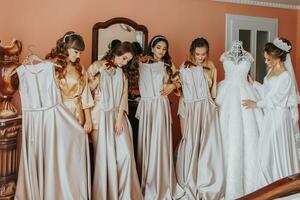 Young bridesmaids in silk robes show off their dresses in a hotel room. Beautiful women celebrating a bachelorette party next to the bride in themed pajamas. photo