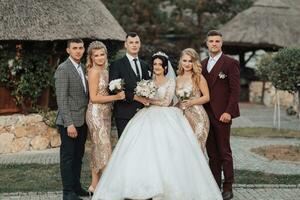 Front view of newlyweds standing and hugging, between friends. Girls in golden dresses hold bouquets, the bride in a voluminous dress, the groom and his friends in suits. Wedding in nature photo