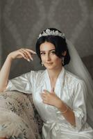 Portrait. A brunette bride in a dressing gown and a chic crown sits on a sofa and poses. Gorgeous make-up and hair. Voluminous veil. Wedding photo. Beautiful bride photo