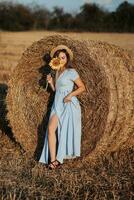 Portrait of a young girl. A girl in a blue dress and a hat holds a sunflower flower covering her face against a background of hay bales. Long straight hair. Nice color. Summer photo