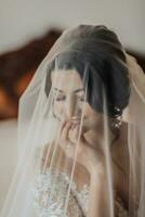 Portrait. A brunette bride in a white dress, wrapped in a veil, looks down and touches her face, poses. Gorgeous make-up and hair. Voluminous veil. Wedding photo. Beautiful bride photo