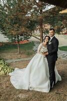 Boda retrato en naturaleza. el morena novia y novio en un blanco largo vestir son en pie y abrazando en contra el antecedentes de conífero arboles y un blanco cabaña. foto