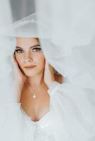 Beautiful curly brown-haired bride in a white dress poses for a photographer, standing under a veil in a beautiful dress with sleeves. Wedding photography, close-up portrait, chic hairstyle. photo