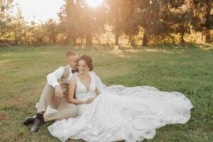 Wedding portrait of the bride and groom. The bride and groom are sitting on the green grass and posing. Behind the bride and groom, the beautiful sun shines through the large Christmas trees. photo