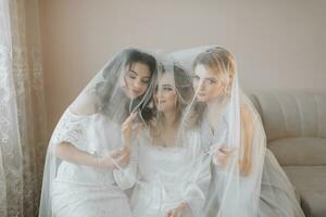 The bridesmaids look at the smiling bride. All on the same couch under a veil. The bride and her fun friends celebrate the bachelorette party in different dresses. Bride and friends in the room photo