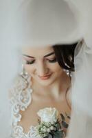 A brunette bride in a white dress holds her wedding bouquet, poses. French manicure. Open shoulders. Beautiful hands. Long veil. Morning of the bride. Details photo