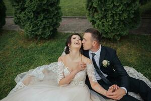 Portrait of the bride in nature. A brunette bride in a white lace dress and a stylish groom in a black suit are sitting on the grass, posing, holding a bouquet. Beautiful makeup. Open shoulders. photo