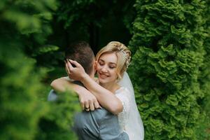 Portrait of the bride and groom standing on the background of green trees, embracing. The bride smiles sincerely. Stylish groom. Fashion and style. Beautiful bride photo