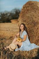 Portrait of a young girl. A girl in a blue dress holds a sunflower flower against a background of hay bales. Long straight hair. Nice color. Summer photo