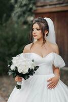 The bride in a voluminous white dress with open shoulders and a long veil, holds a bouquet of white roses and looks to the side. Portrait of the bride. Beautiful makeup. Wedding in nature photo