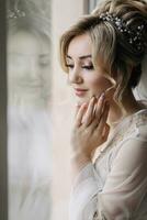 Blonde bride with elegant hairdo and tiara, wearing a robe, posing, looking at the camera, touching her face. Beautiful makeup. Elegant hairstyle. French manicure photo