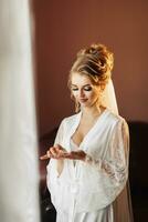 A blonde bride in a long, lacy petticoat is standing in her room, posing by the window, holding her earrings. Beautiful hair and makeup, open bust. photo