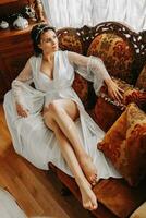 hermosa novia en un retro sofá en un túnica con abierto piernas, lleno longitud foto. Boda peinado, ligero maquillaje foto