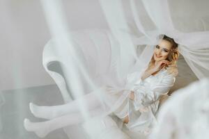 Blonde bride wearing a satin robe and lying on a couch, posing and showing off her tiara and veil. Beautiful hair and make-up, open bust. Wedding portrait. Sincere smile photo